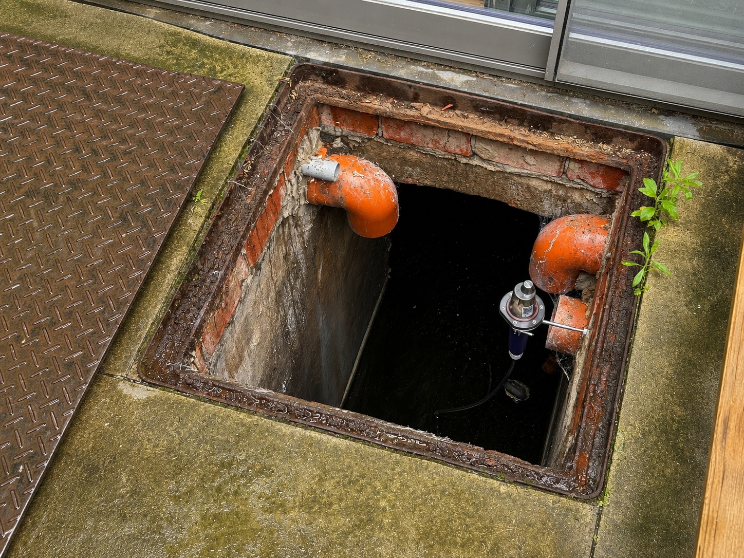 Blik in een open regenwaterput naast een woning: gemetselde schacht met twee oranje aansluitbochten en een niveausensor aan een beugel boven het water