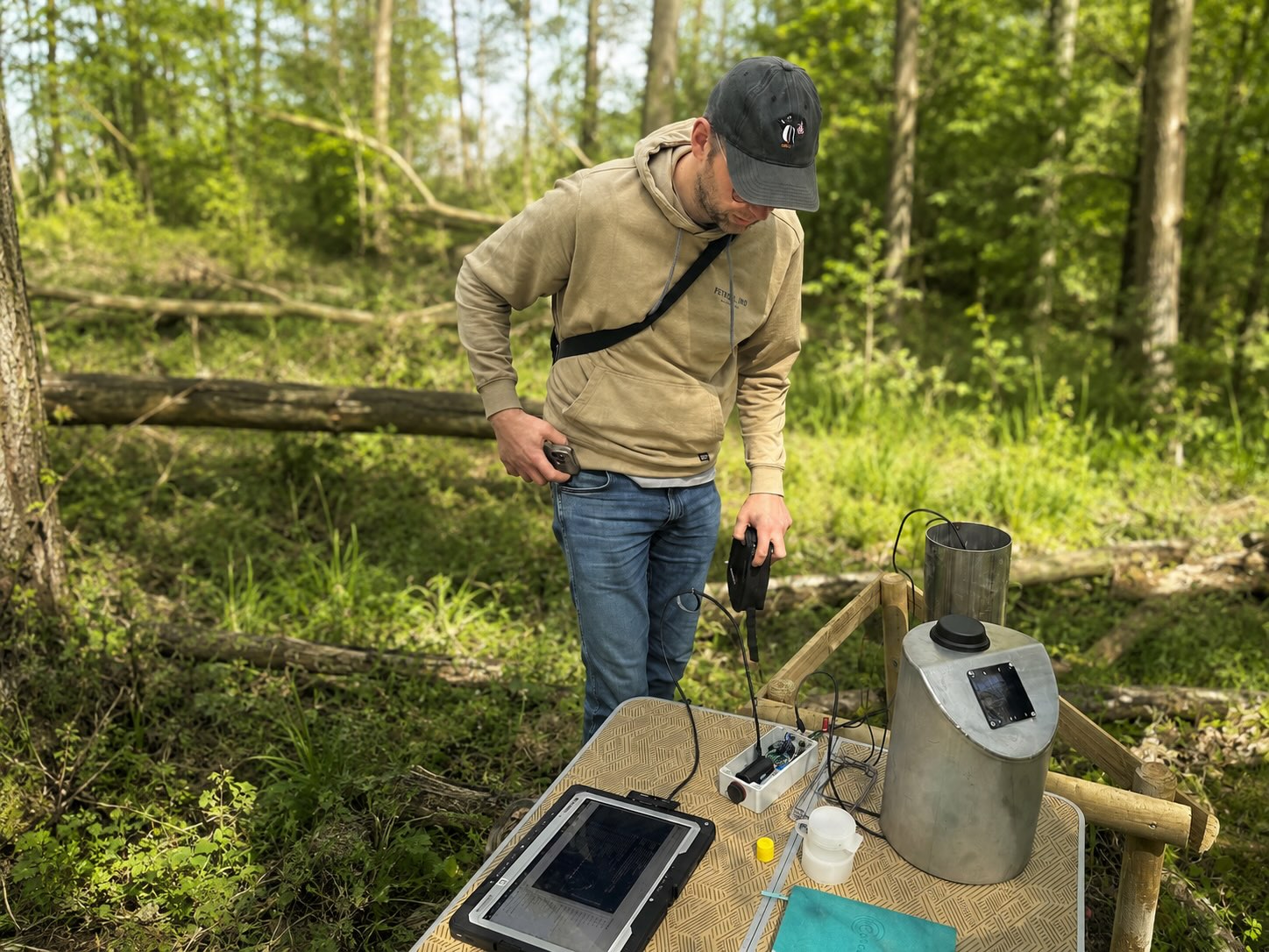 Medewerker bij een grondwatermeetpunt in een bos, met een veldtablet en een geopende IoT-module op een werktafel