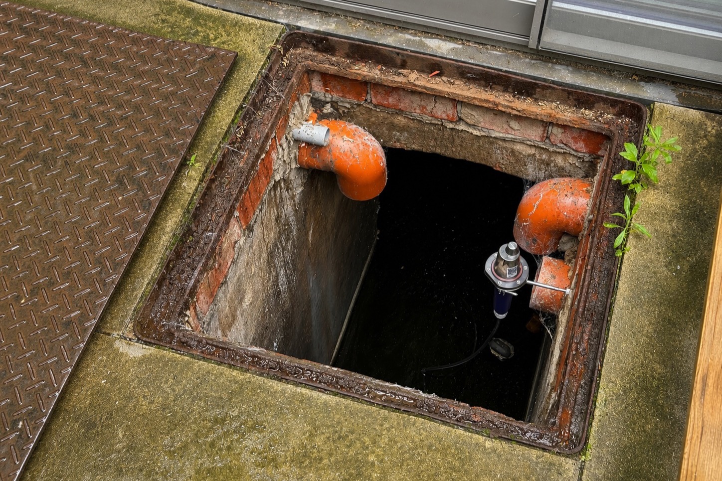 Open regenwaterput naast een woning, met twee oranje aansluitbochten en een niveausensor boven het water